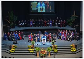 Item is an aerial photograph of President Neil Snider standing at a podium speaking at the 49th Annual Commencement Ceremony held in the auditorium of Abbotsford Pentecostal Assembly in Abbotsford, BC. Seated on stage are Board of Governors, and faculty members. The ceremony was projected on an oversized screen that is seen on the top of the photograph.