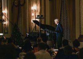 Item is a photograph of President Bob Kuhn speaking on stage during the Christmas in the City fundraising event. The photograph was take on angle from the right side and there is a grande piano in the background. This event was held in a ballroom at the Fairmont Hotel in Vancouver.